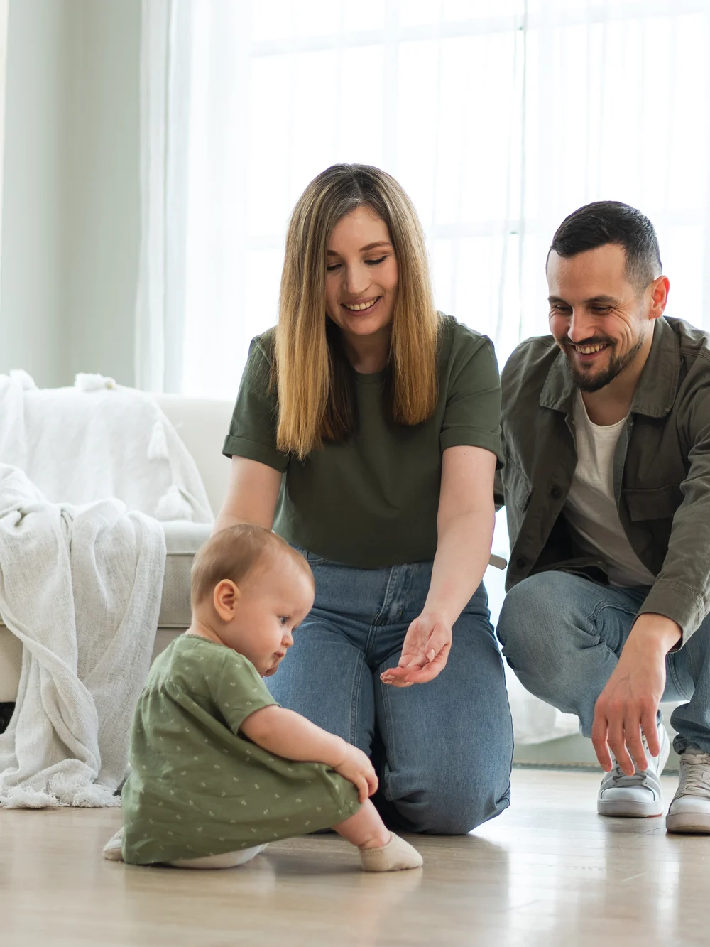 Two parents crouch on a living room floor near a baby sitting a short distance away