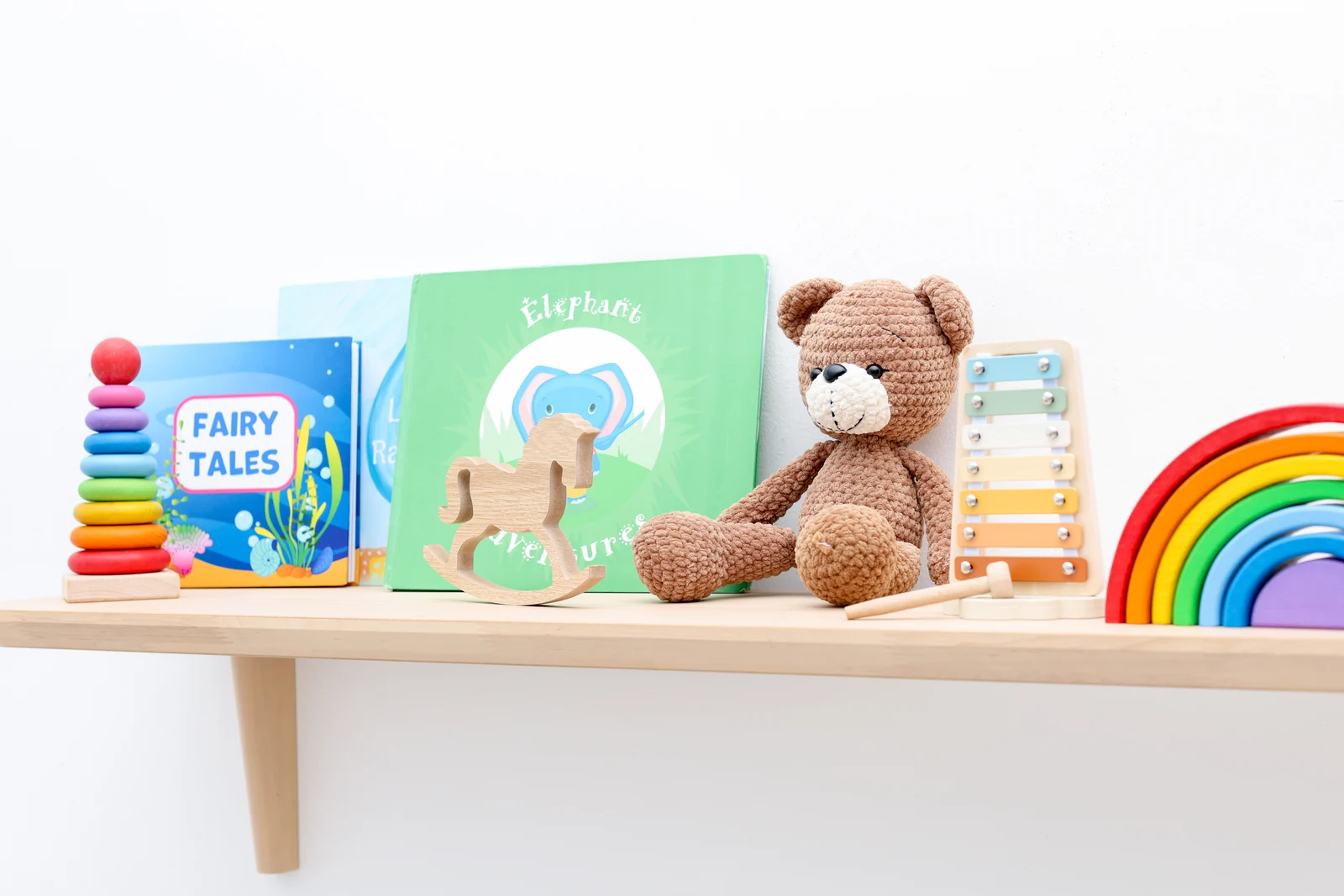 A wall shelf with board books, a stacking ring toy, a teddy bear and a wooden rainbow