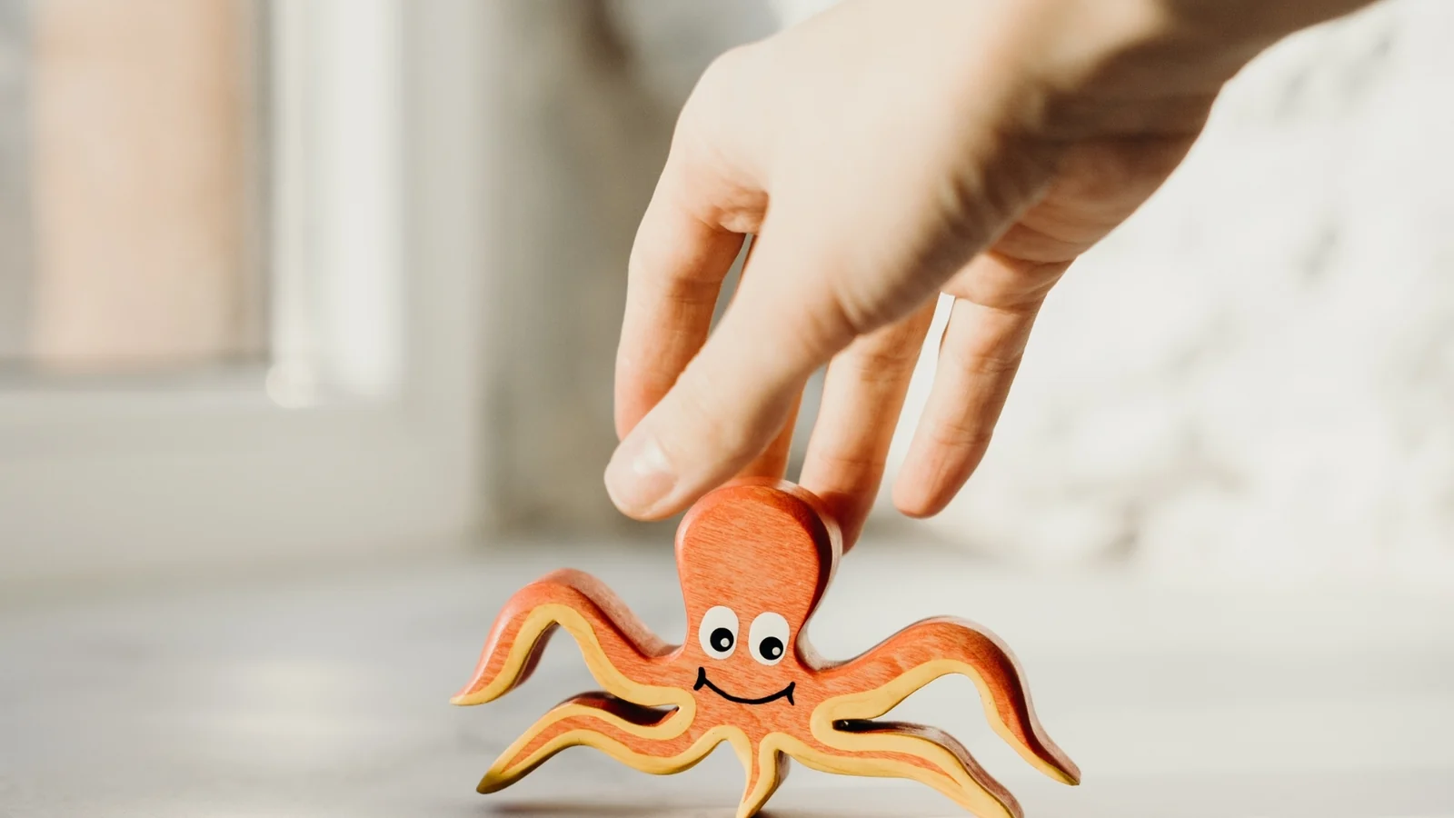 A hand lifts a small painted wooden octopus toy from a sunlit table, checking what makes a toy Montessori
