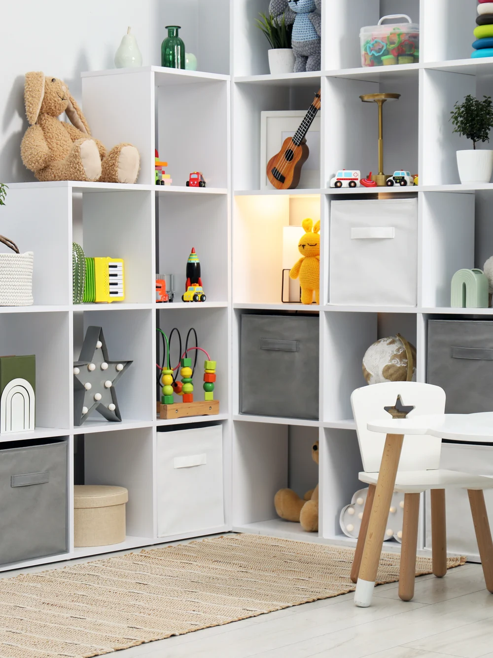 A white corner cube shelf with toys, plush animals and fabric bins, with a small kids' chair and a woven rug in front