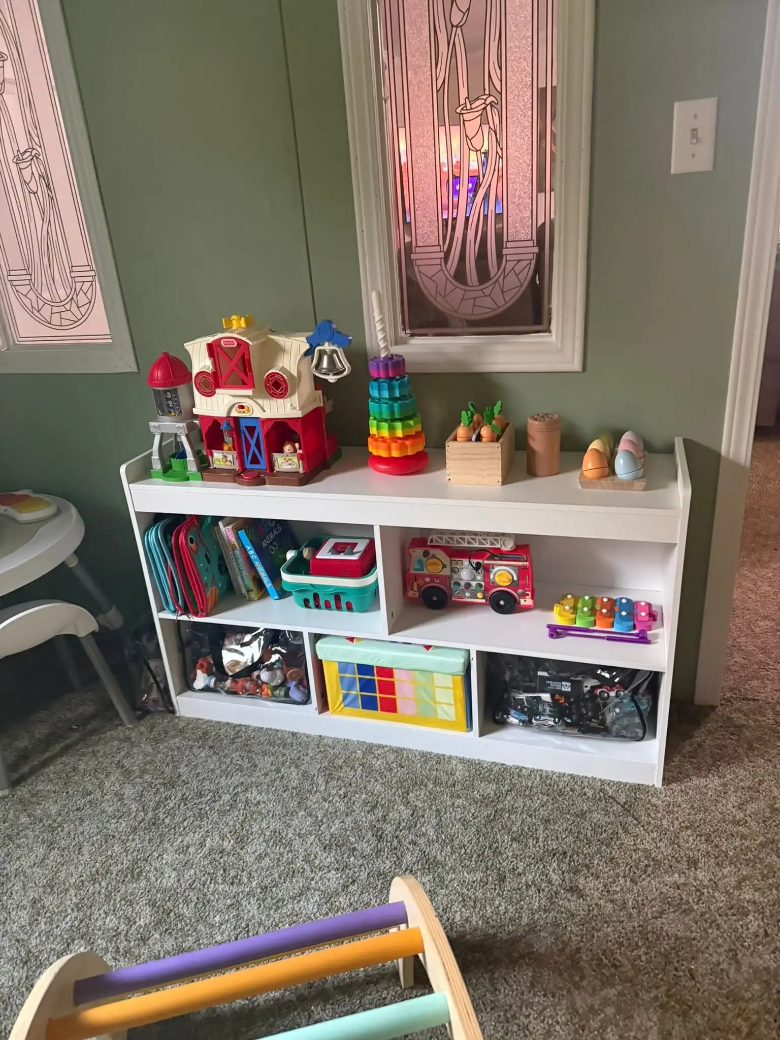 A white open toy shelf with plastic toys, books and a wooden rainbow climber on a gray carpet