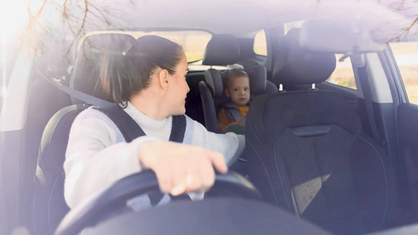 Woman driving with one hand on the wheel, glancing back at a toddler in a car seat in the back seat