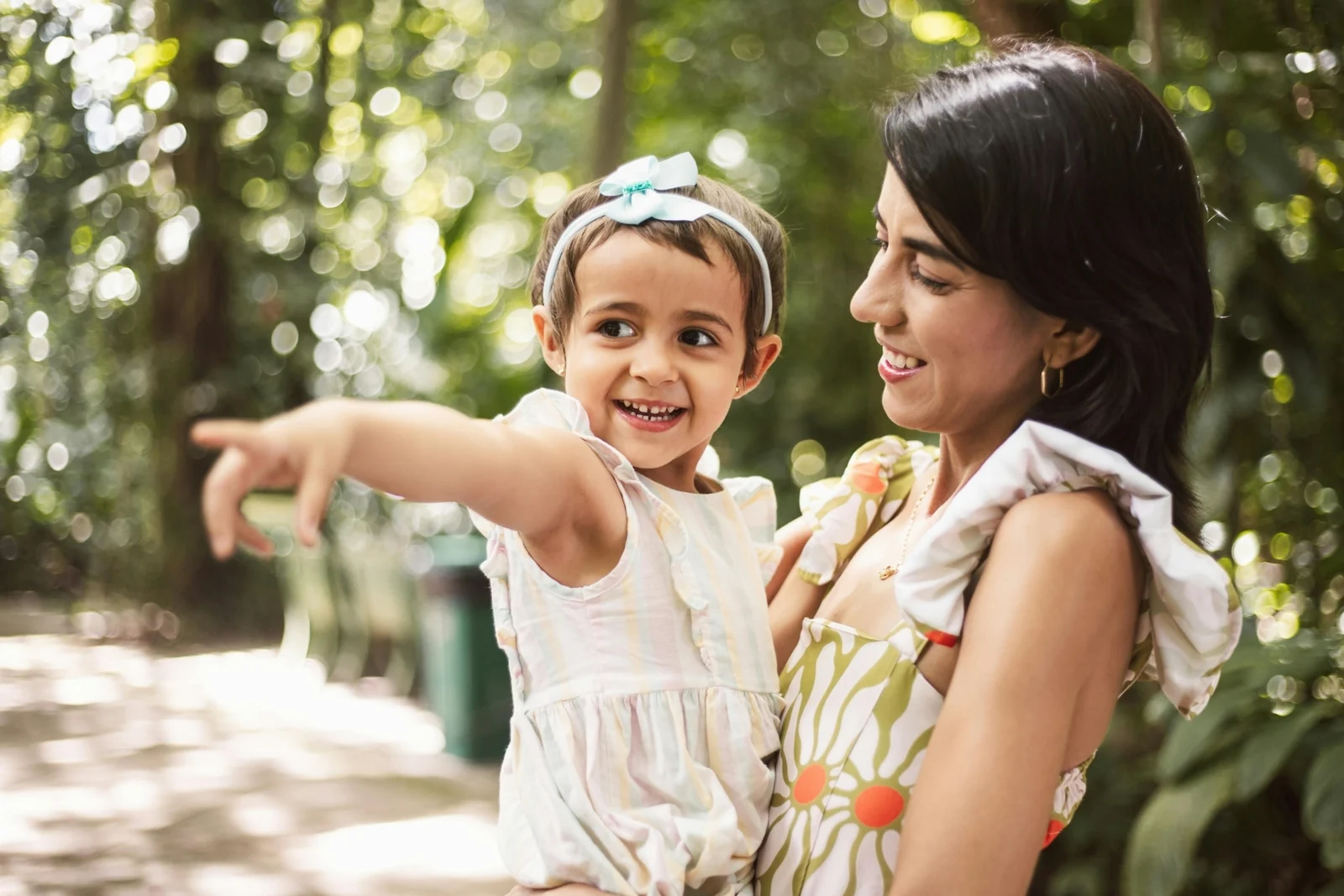 Woman holding a smiling toddler on her arm in a wooded park while the child points at something off to the side
