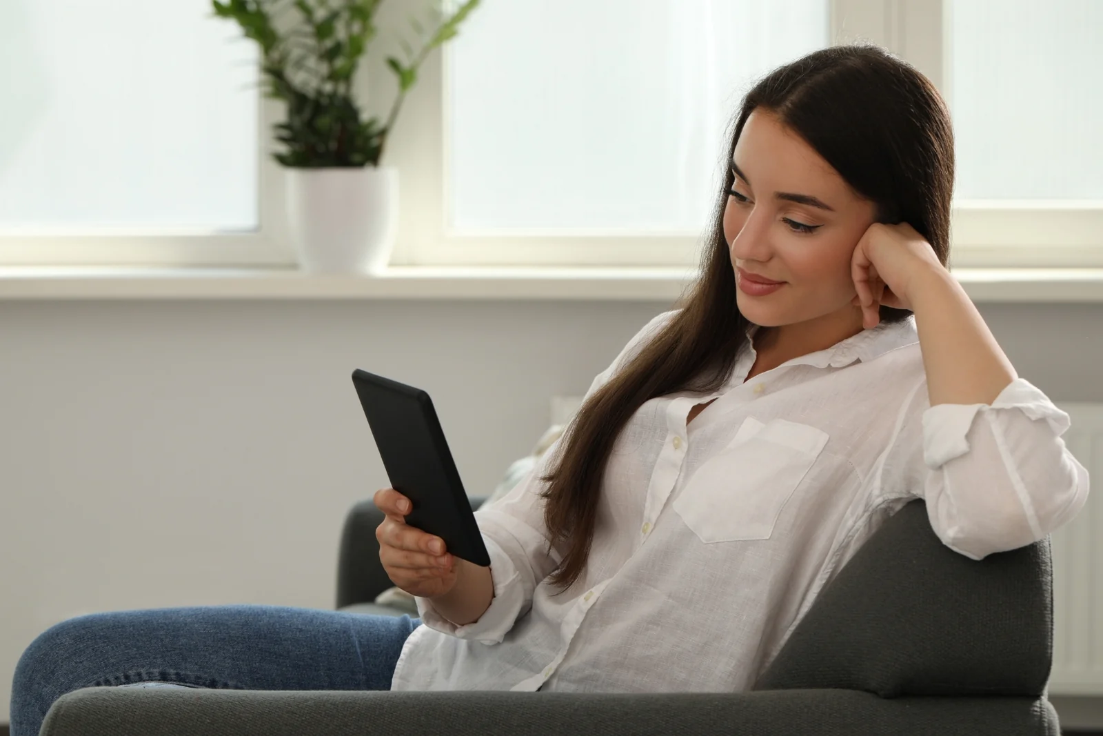 A woman reads on an e-reader in an armchair by a window