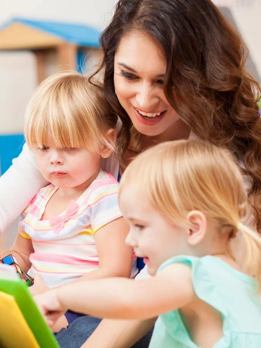 Woman reading a colorful picture book with two toddler girls who are pointing at the pictures