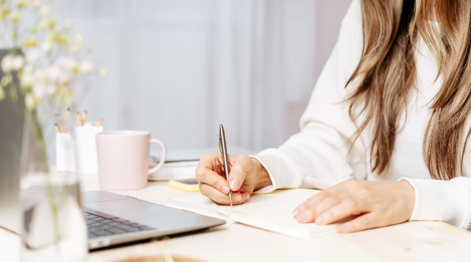 A woman writes in an open notebook at a table, with a laptop and a pink mug beside her