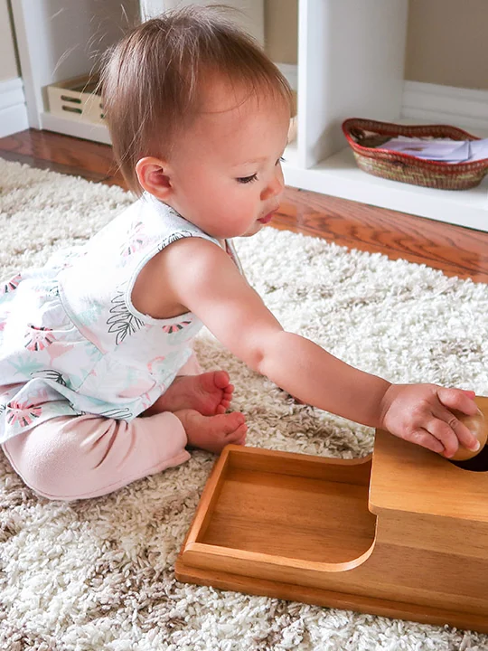 A baby on a shaggy rug pushes a ball into a wooden drop box with a round hole on top