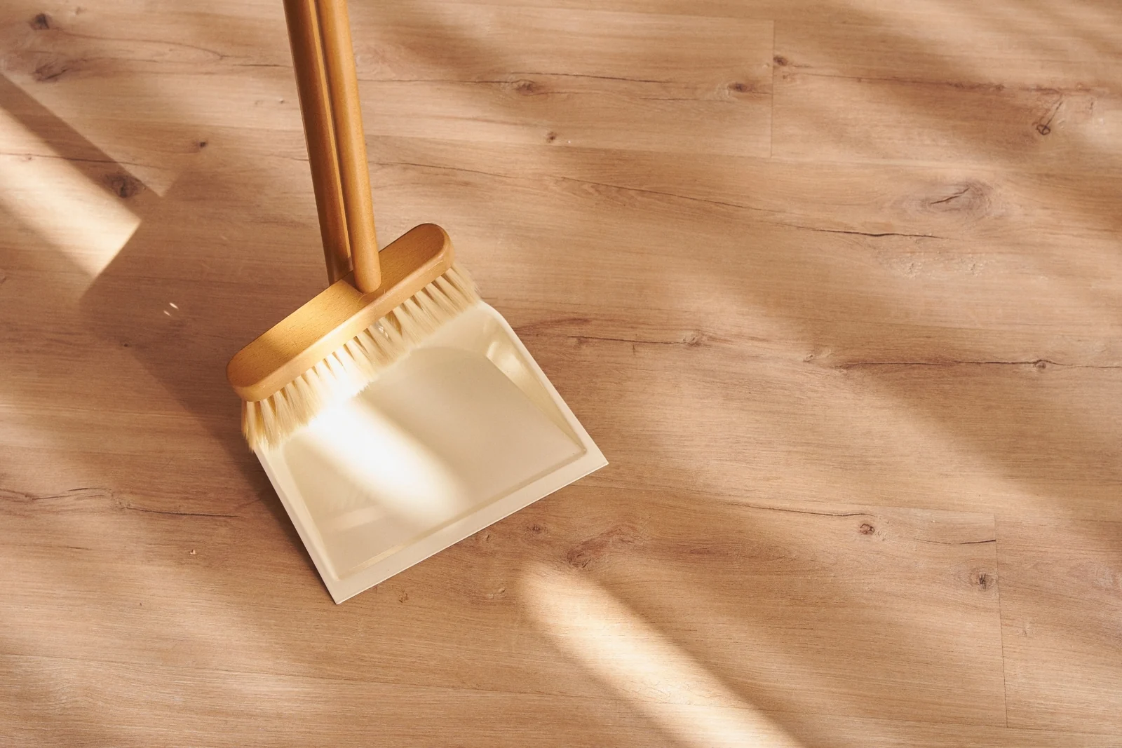 A wooden-handled brush and a cream dustpan standing on a light wood floor in warm sunlight
