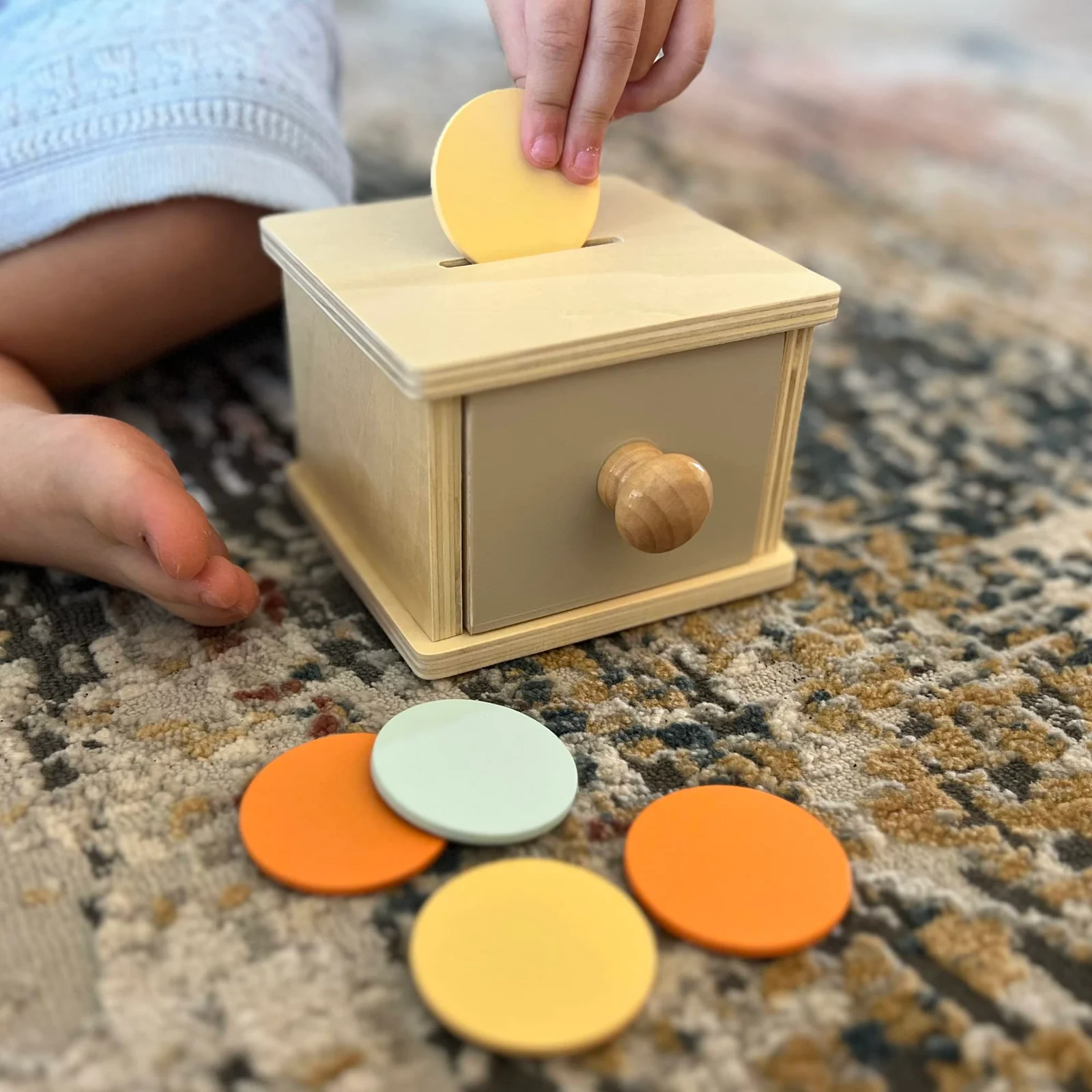 A light wooden coin box with a drawer, a hand dropping a round disc into the slot, and four colorful discs on a rug