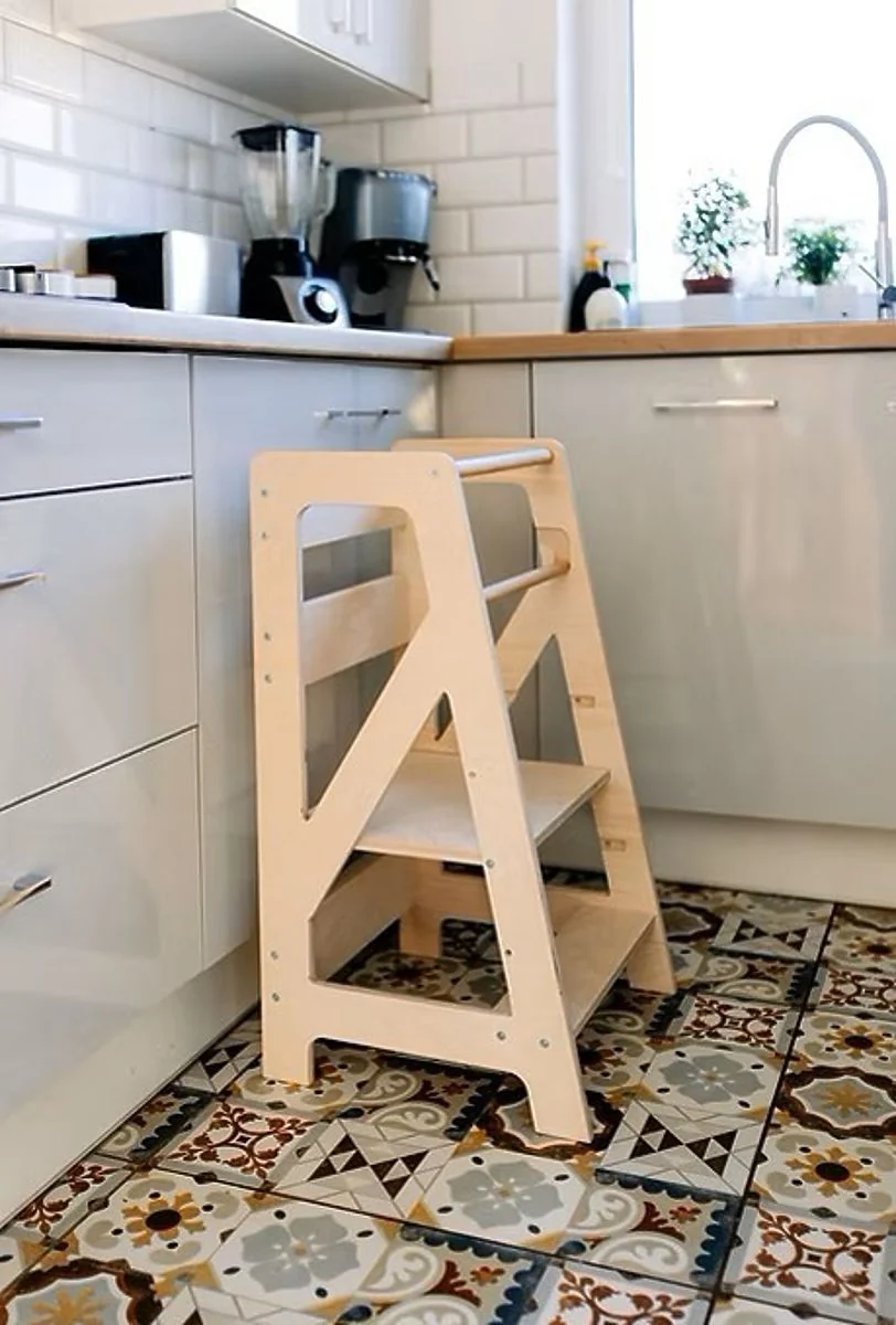Empty wooden learning tower with a railing and steps standing against kitchen cabinets on a patterned tile floor
