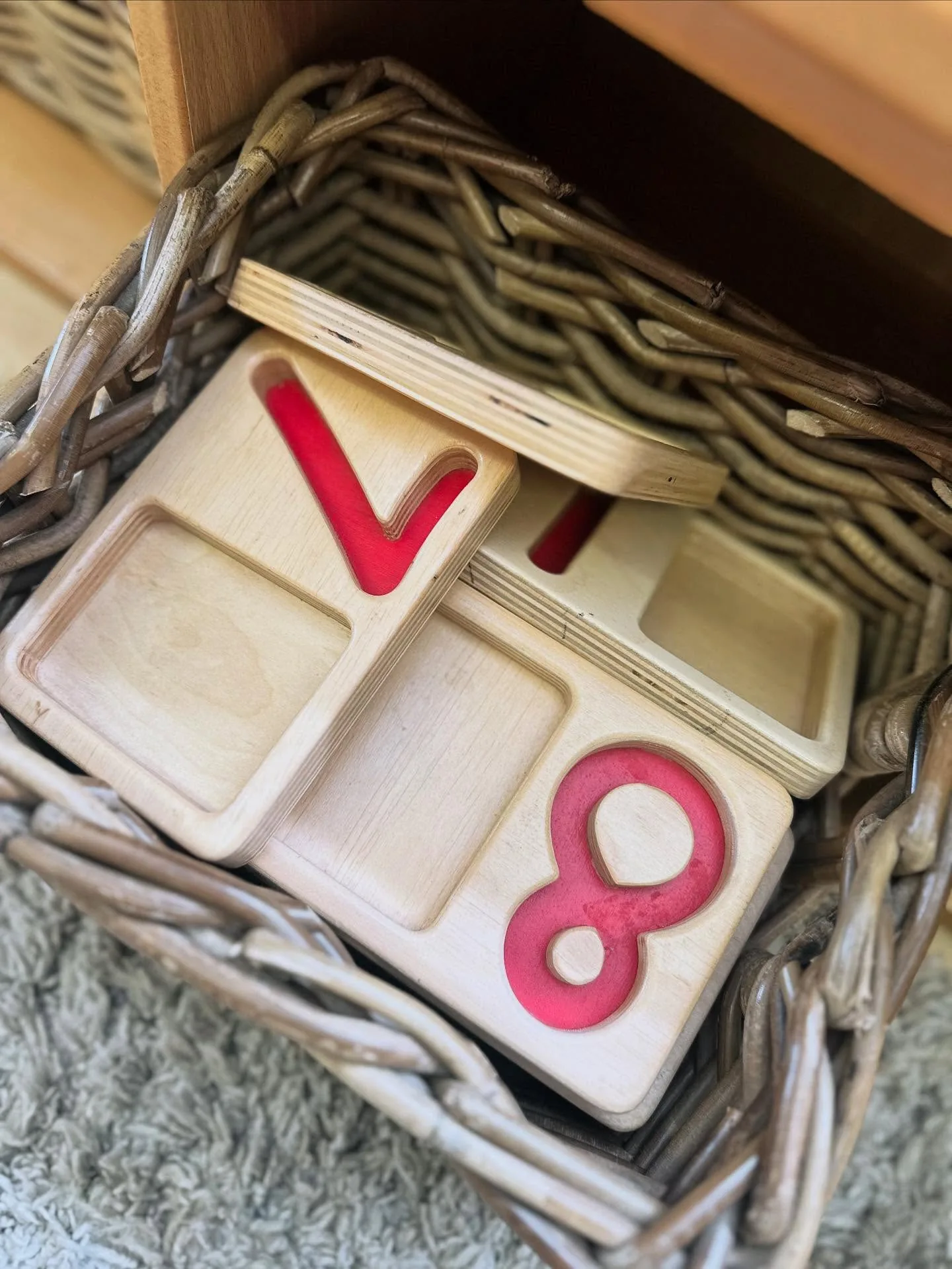 Woven basket on a low shelf holding a stack of wooden number trays with red inlaid numerals, above a gray rug