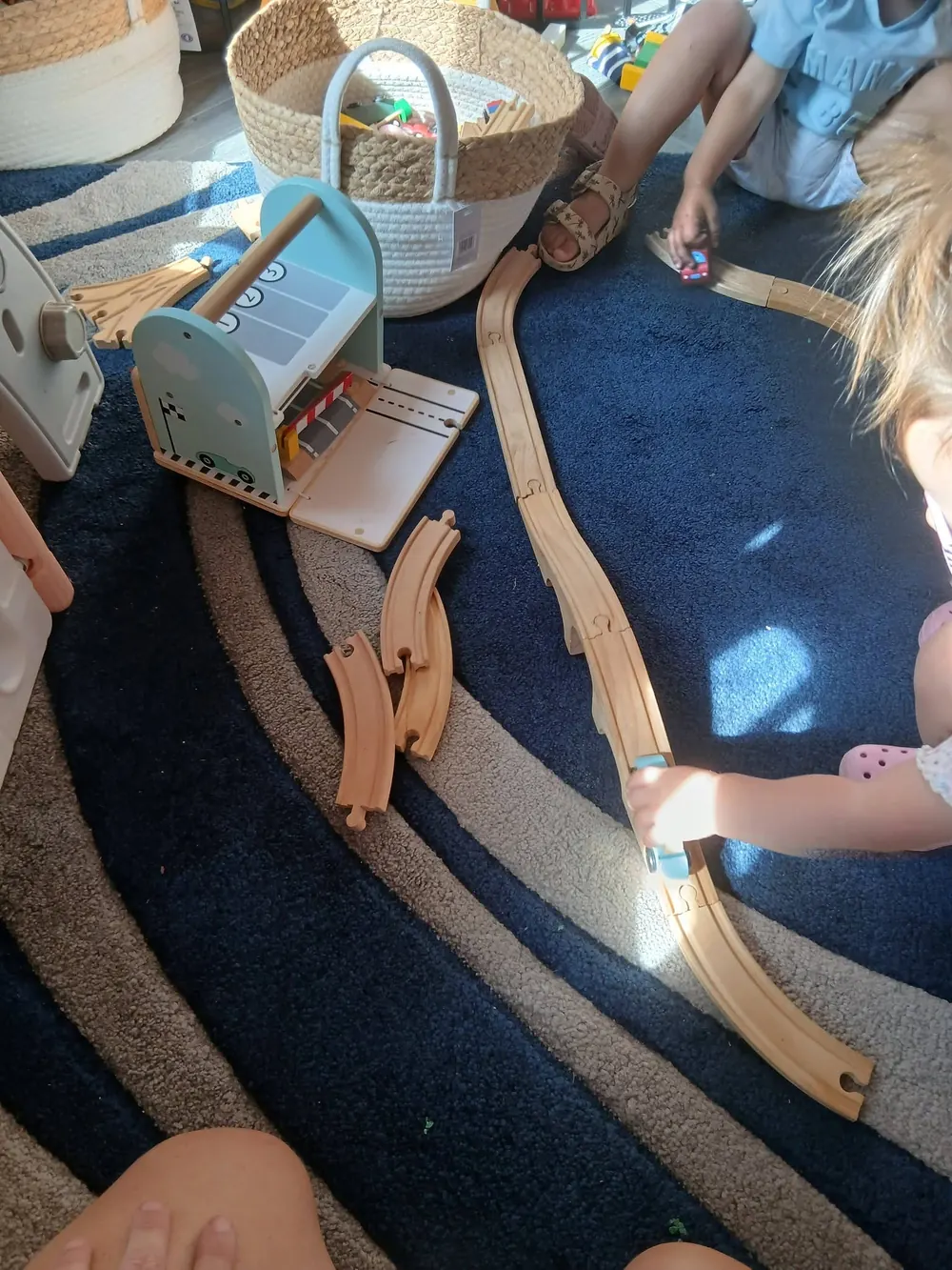 Wooden train tracks and a toy garage spread over a navy rug, with two children playing nearby